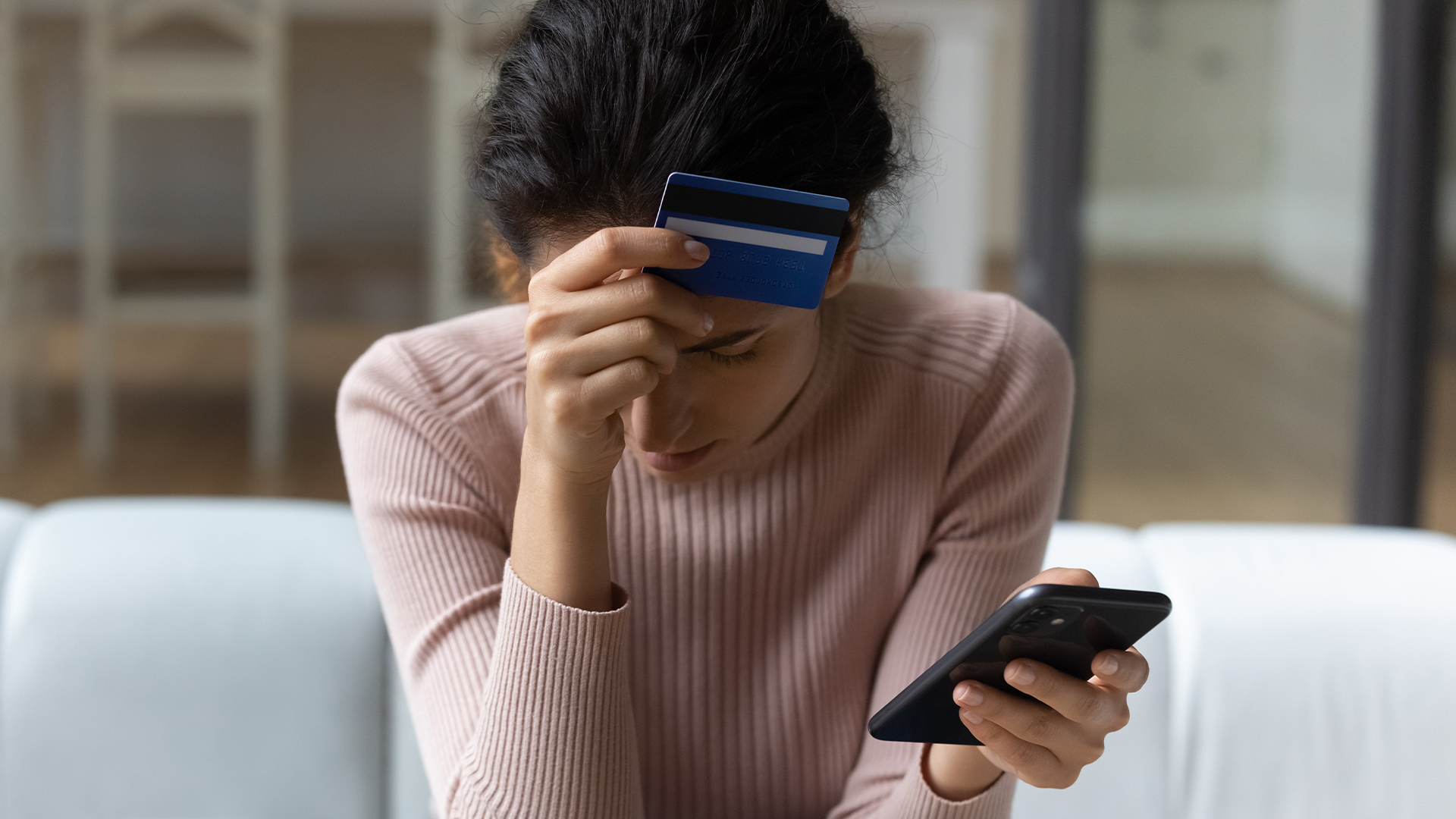 A person sits on a sofa, holding their forehead with one hand and a credit card against their head, while looking at a smartphone in their other hand.